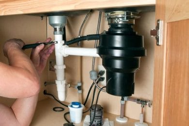 Plumber repairing a garbage disposal beneath a kitchen sink in Englewood